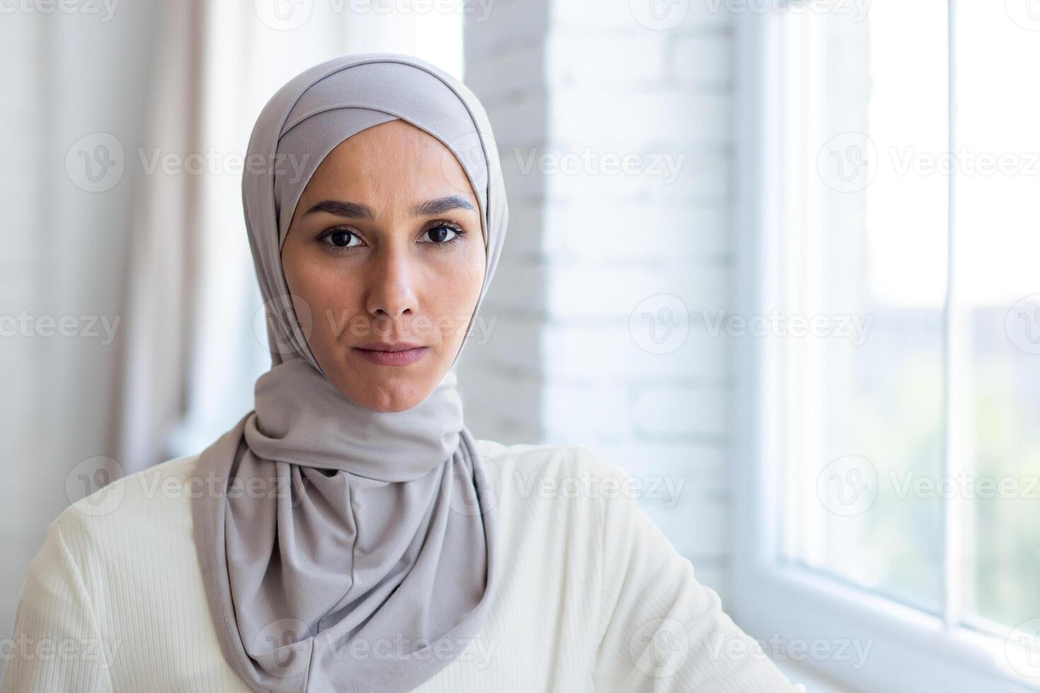 Close-up photo. Portrait of a young Muslim woman in a hijab standing by the window at home, confidently and seriously looking at the camera. photo