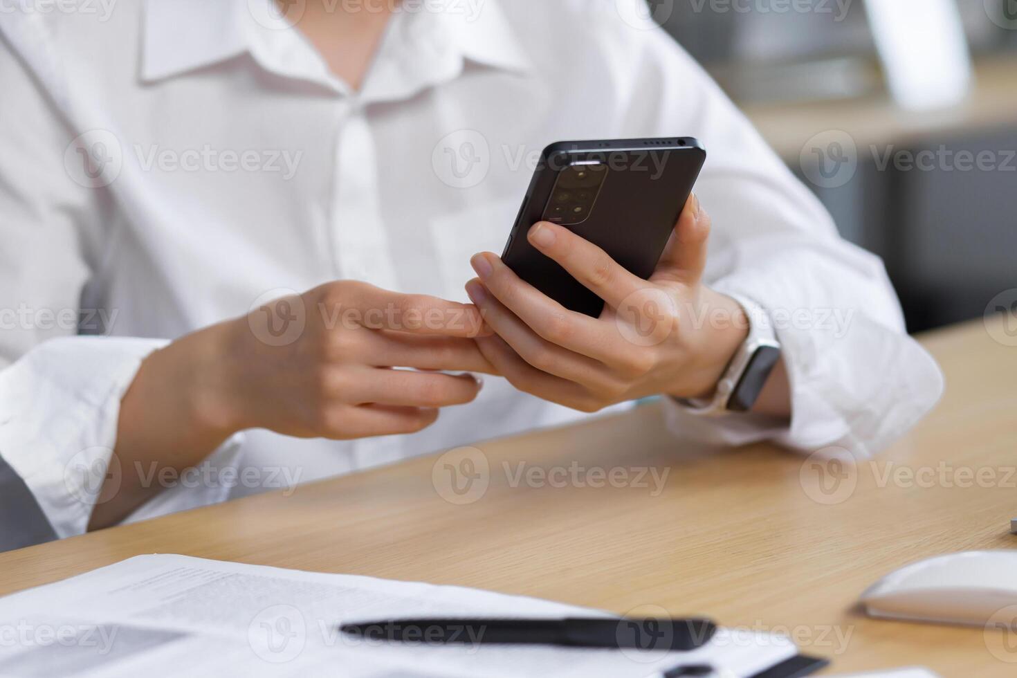 Close-up photo. The hands of a young woman in a white shirt are holding a mobile phone, typing a message at the table. photo