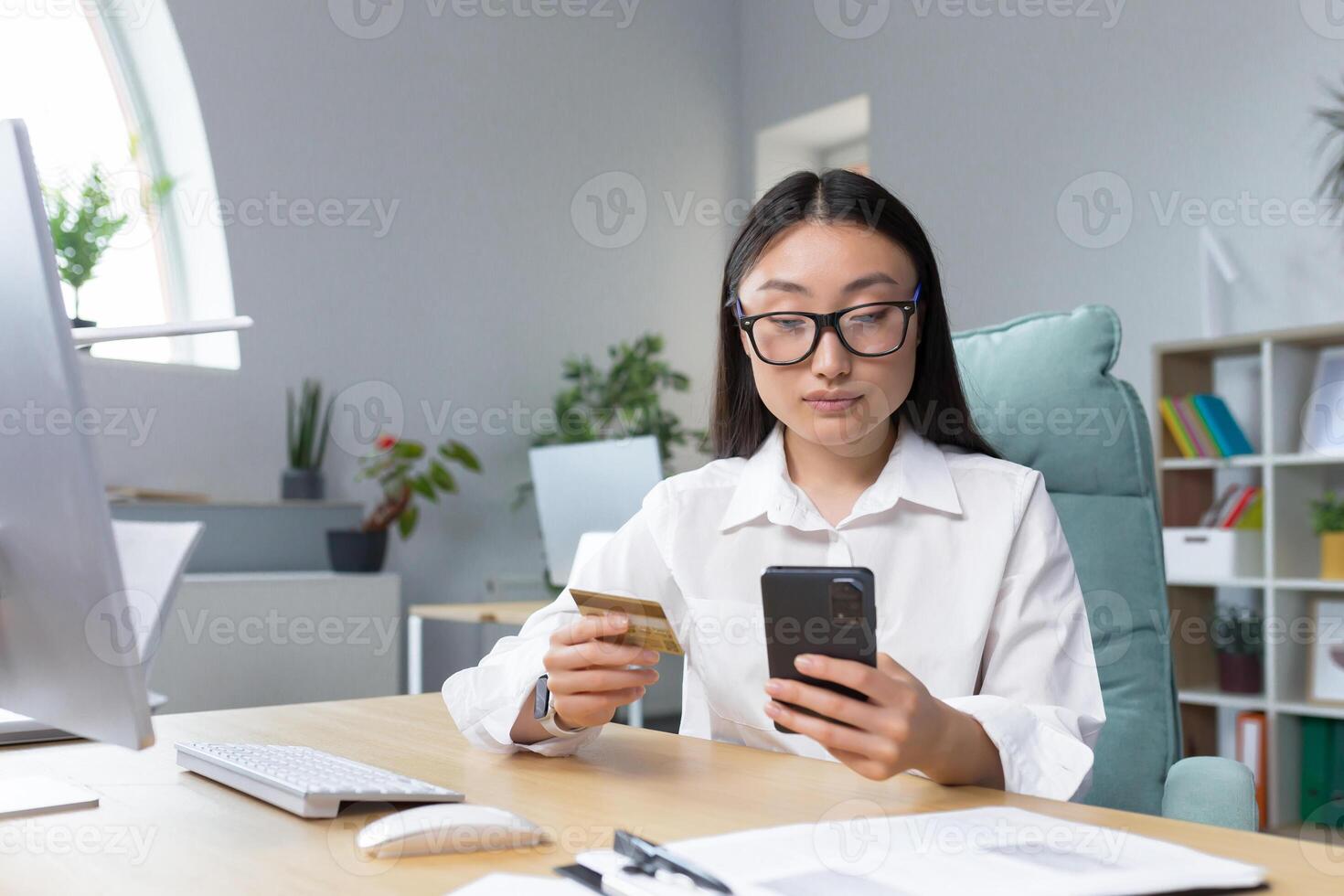 Online shopping. A young beautiful Asian woman holds a mobile phone and a credit card in her hand, makes an order on the phone, pays by card.. Sitting at a table in a modern office. photo