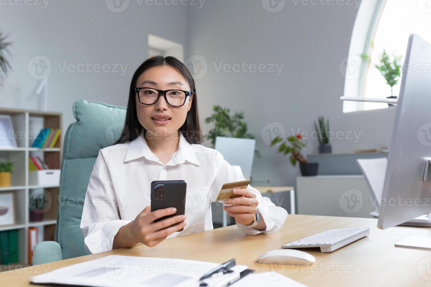 Online shopping. A young beautiful Asian woman holds a mobile phone and a credit card in her hand, makes an order on the phone, pays by card.. Sitting at a table in a modern office. Looks into the camera. photo