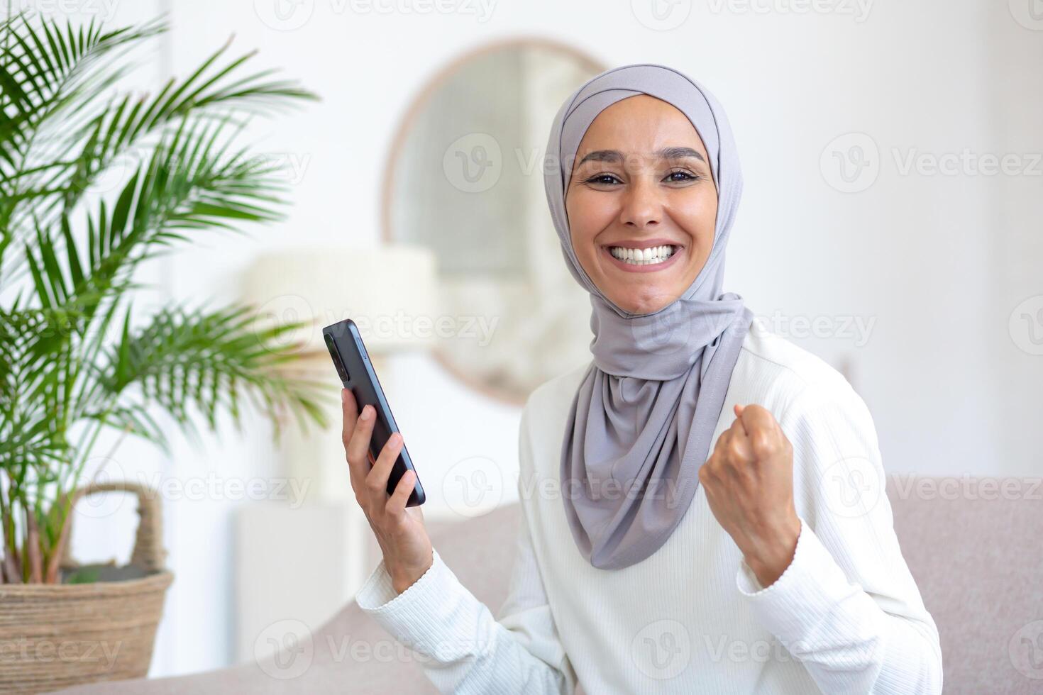 Happy young female Muslim student in hijab is happy and celebrating on camera. shows a victory gesture. holds the phone in his hands. received good news, message, e-mail. photo