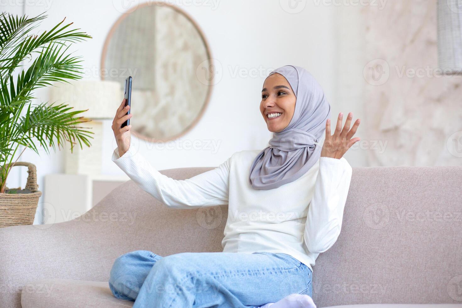Smiling young Arab woman in hijab sitting at home. in the hotel on the sofa and talking on the phone on a call. photo