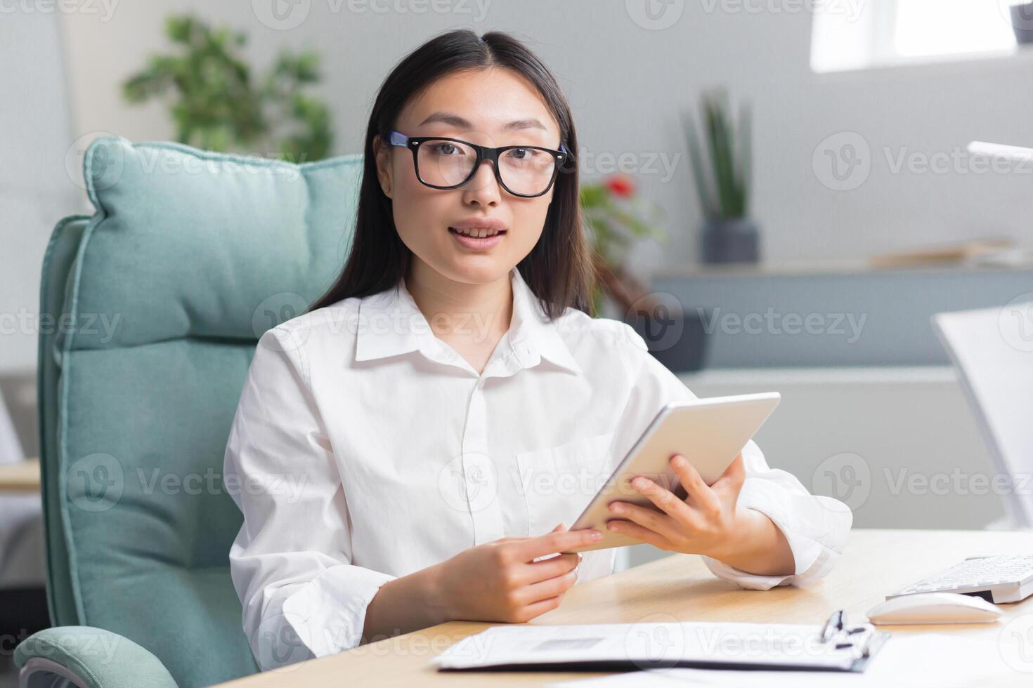Young beautiful Asian business woman in glasses sits in the office at a computer desk. Holds a tablet in her hands, types, works. Looks at the camera, smiles. photo