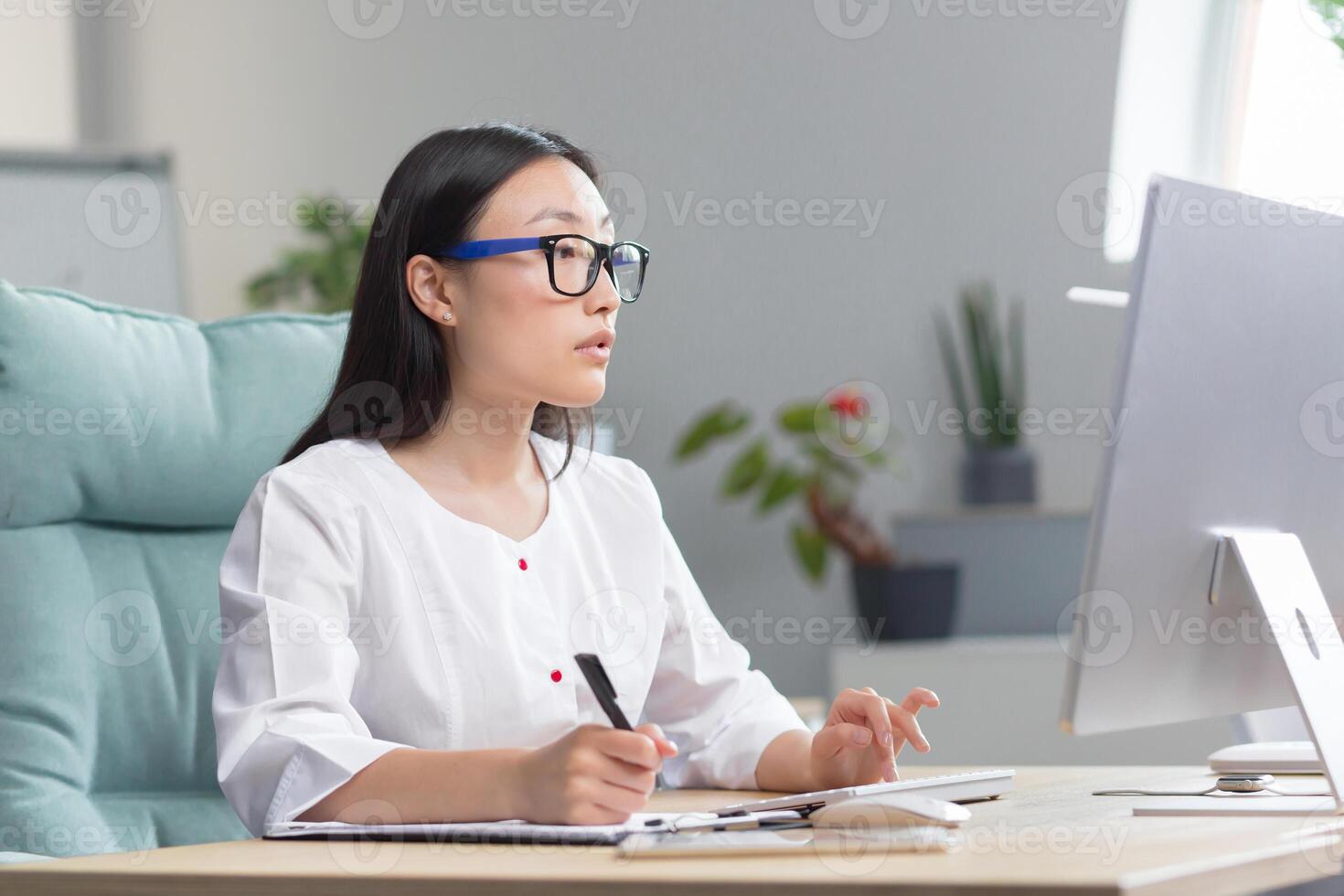 The doctor is online. A young beautiful Asian woman doctor conducts an online consultation. He is sitting in a modern office at a work table, talking on a call with a patient at a computer. photo