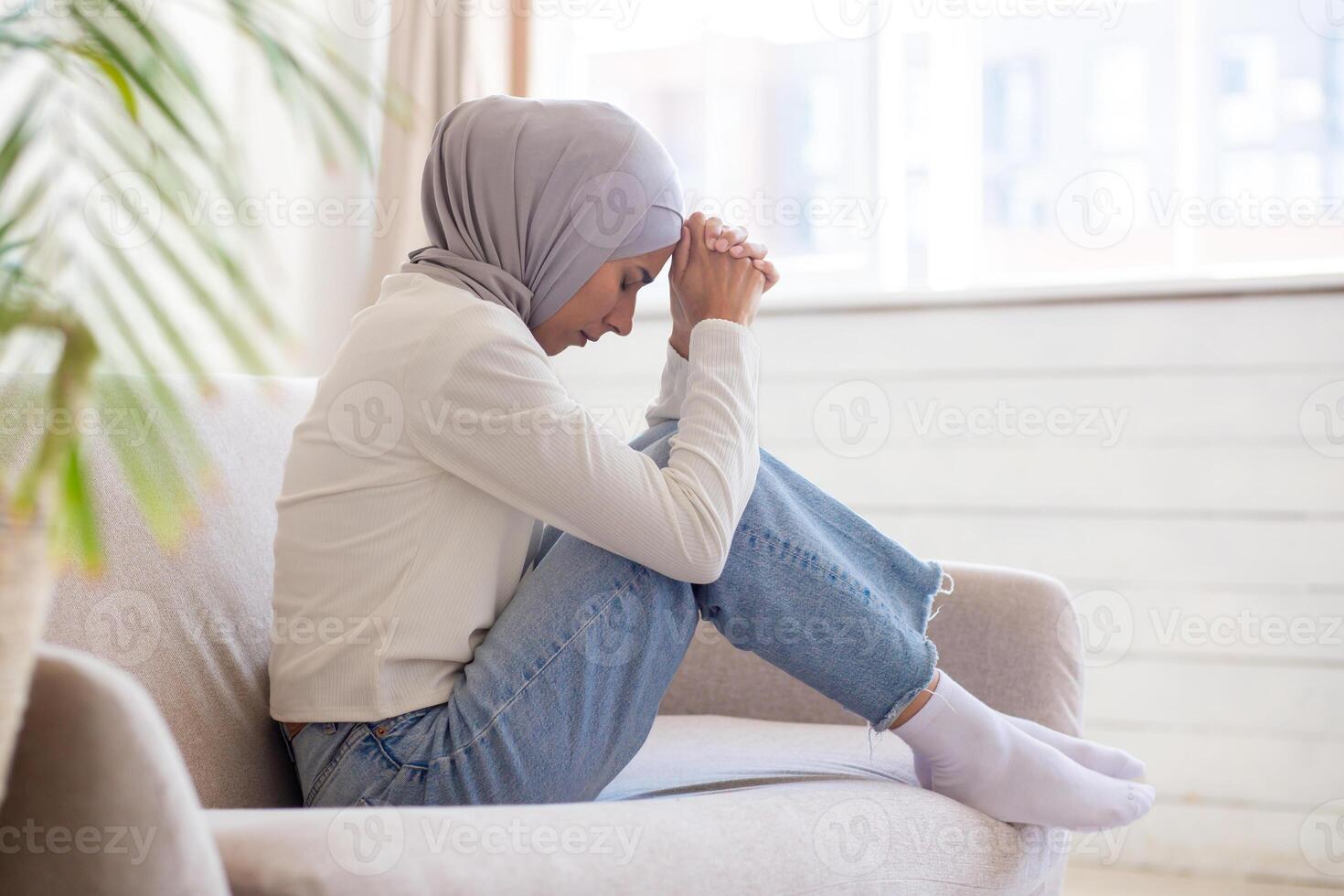A young Muslim woman in a hijab sits cross-legged on the sofa, resting her head thoughtfully on her hands, experiencing depression, stress, loss. divorce, war. refugee She closed her eyes and prayed. photo
