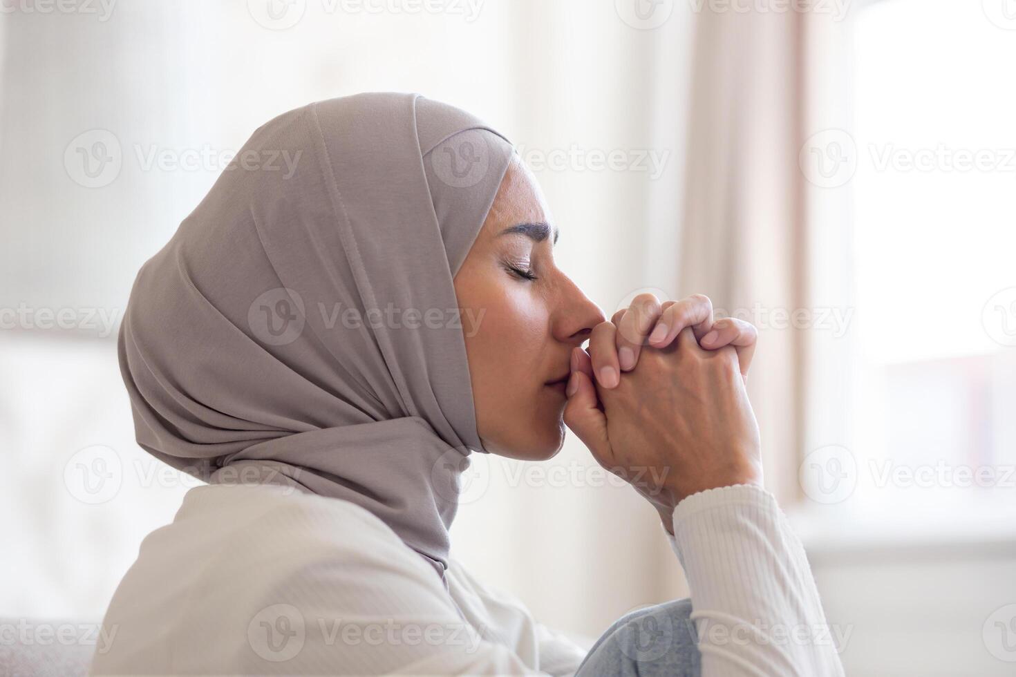 Close-up photo. Portrait of a young Arab woman in a hijab at home, who folded her hands together, closed her eyes and prays, suffers from depression, thinks about solving problems. photo