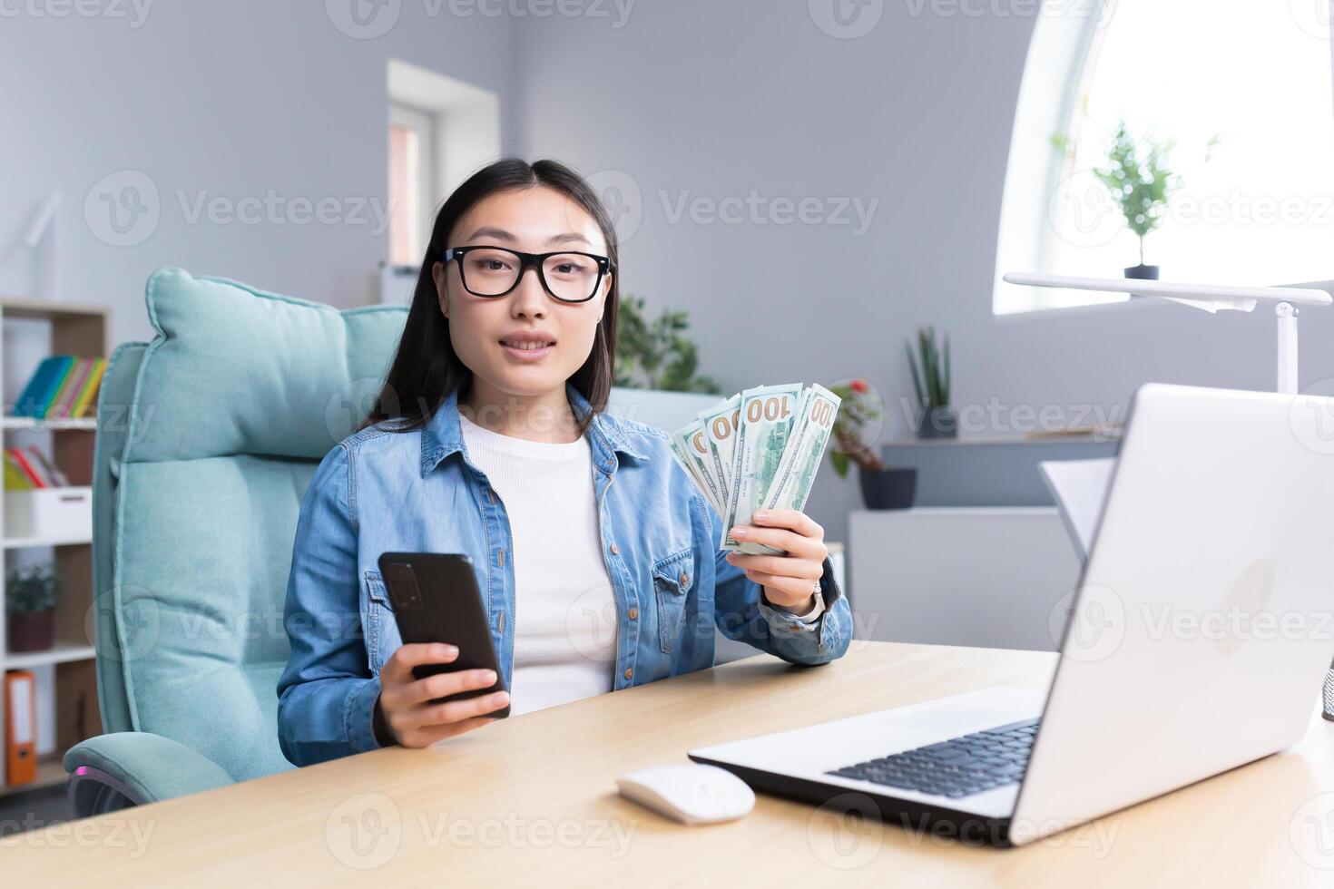 Business training about money. A young Asian business woman conducts a training session on the topic of money. He is holding a phone and a wad of bills. Sits at his desk, talks, looks into the camera. photo