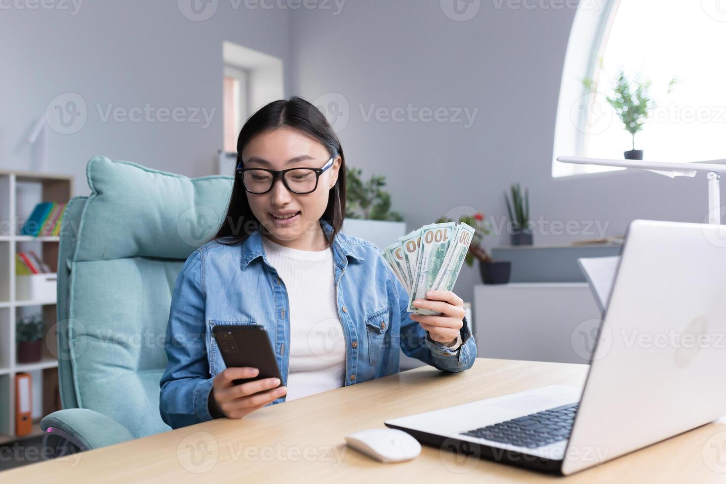 Successful Asian business woman is happy with winning in online casino, looking at phone screen and holding money dollars in hands, business woman working in office. photo
