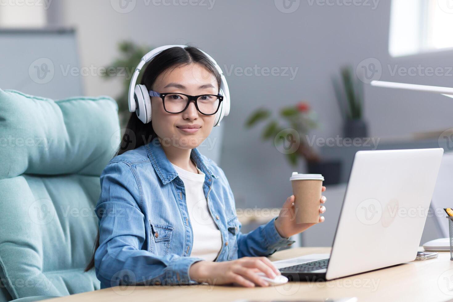 Online training. Young beautiful Asian student girl in glasses studies at a laptop in headphones, drinks coffee. Learns remotely, sits at a desk in a modern office, looks at the camera. photo