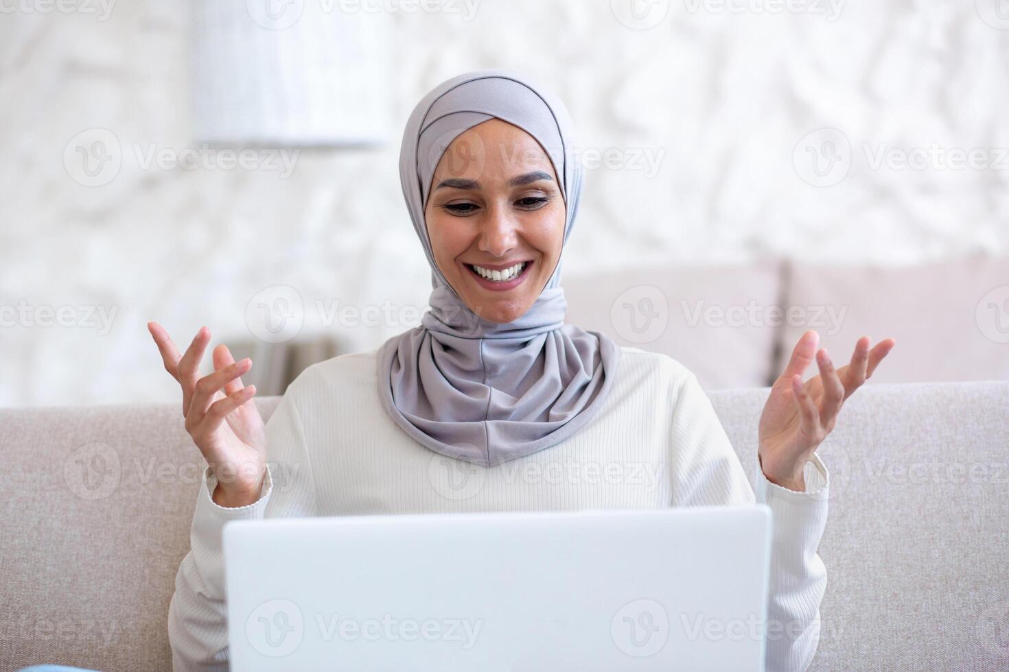 Close-up photo. Muslim young woman in light hijab talking on laptop call at home. He smiles, greets, gestures with his hands. photo