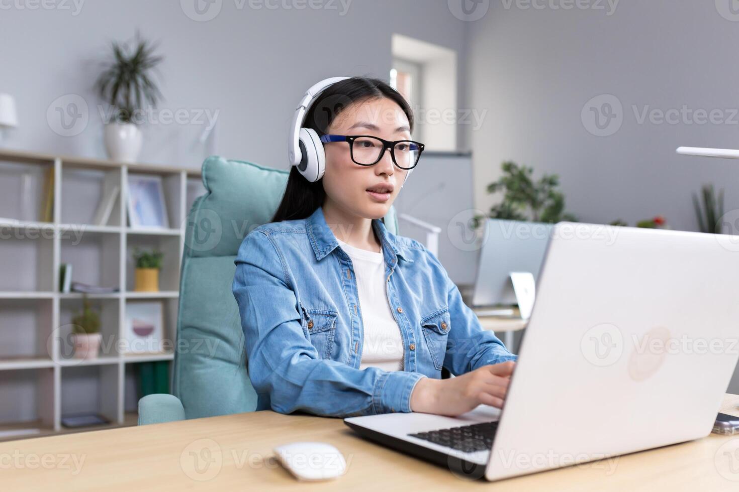 Online training. Young beautiful Asian girl student in glasses studying at a laptop in headphones. Learns remotely, sits at a desk in a modern office. photo