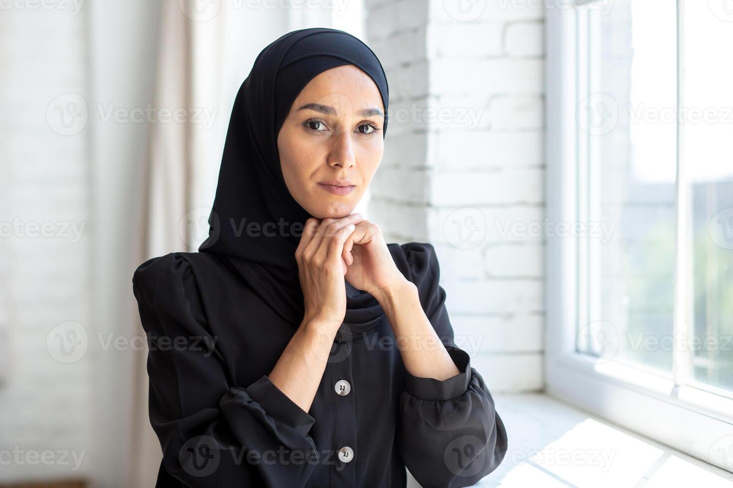 Portrait of a young Muslim woman in a black hijab standing indoors by the window, folding her arms and posing and seriously looking at the camera. photo