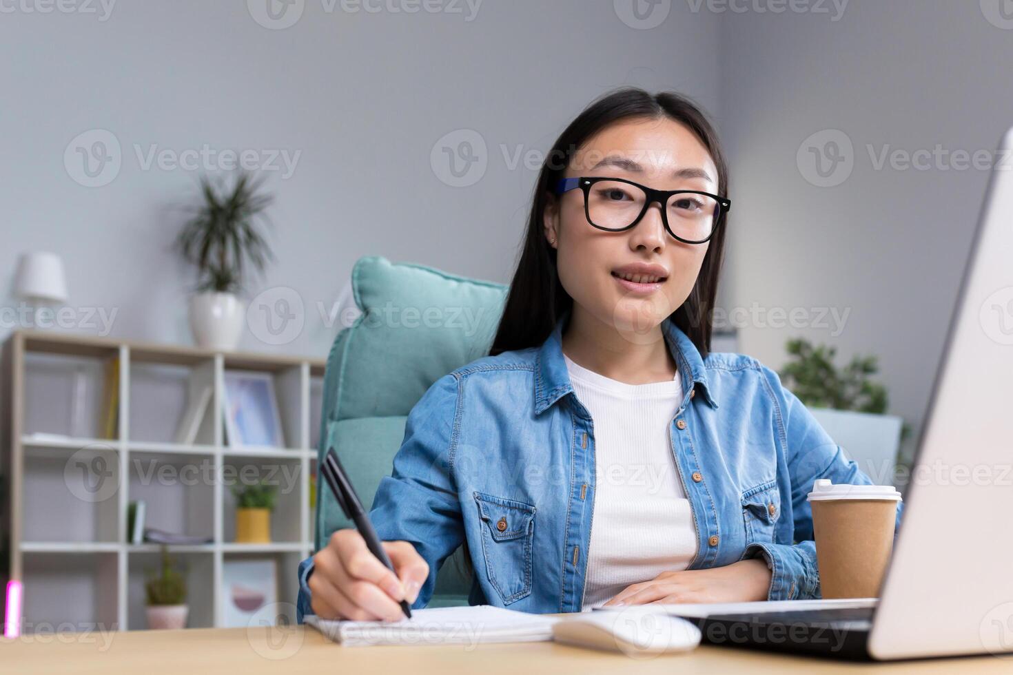 Job interview. A young beautiful Asian business woman in glasses sits at a desk, conducts an interview in the office, hires, writes down a questionnaire in a notebook, interviews the interlocutor. Hello, looking at the camera. photo