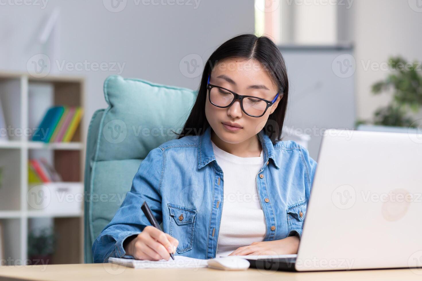 Distance Learning. Young beautiful Asian female student taking an online exam. Sitting in glasses and jeans at the table at the computer, making notes in a notebook. photo