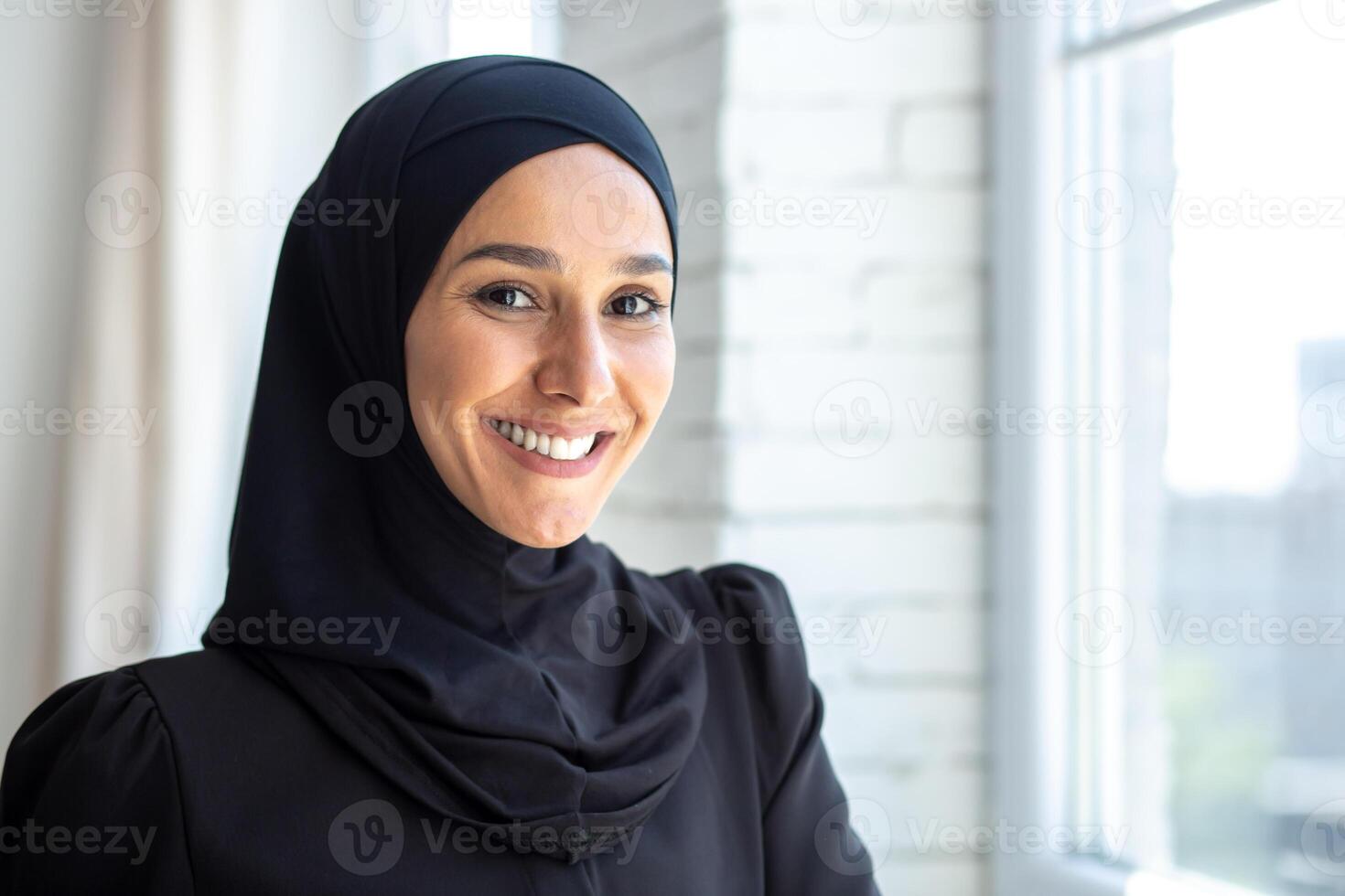 Close-up photo. Portrait of a young beautiful Muslim woman in a black hijab looking confidently and smiling at the camera, standing indoors by the window. photo