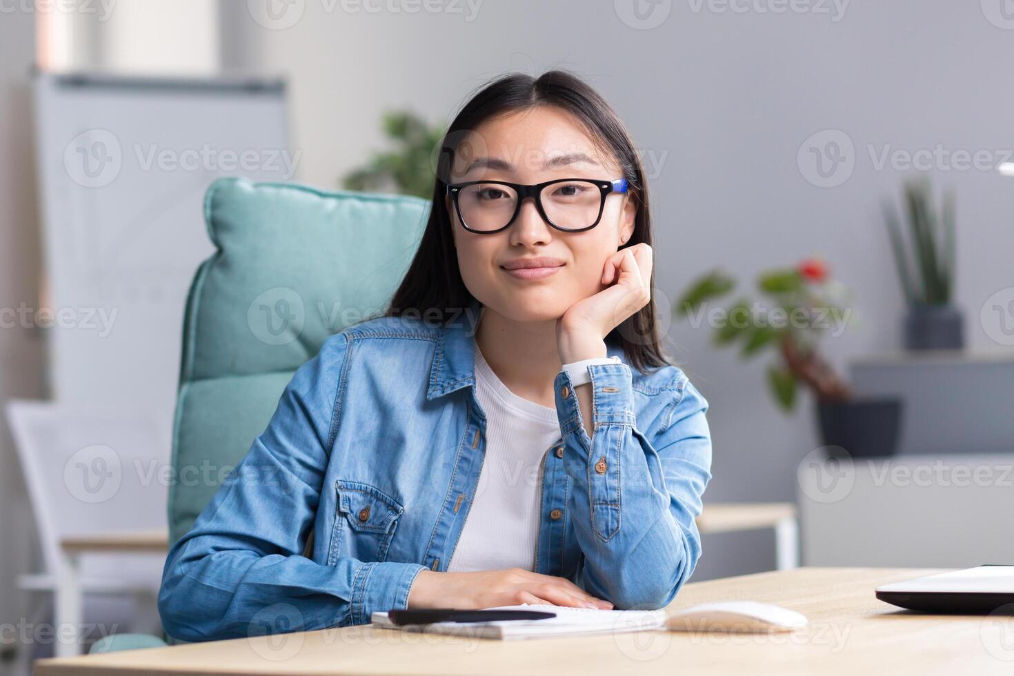Portrait of a young Asian student girl in glasses and denim clothes, sitting at a desk with a notebook and a pen, looking at the camera, smiling. photo