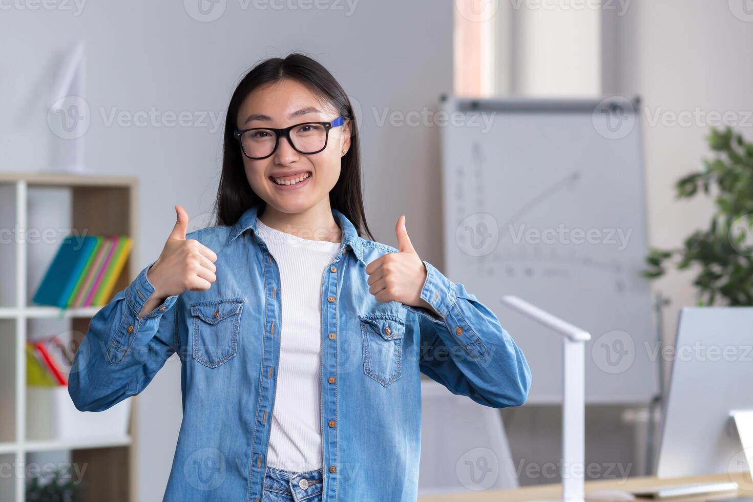 Portrait of young asian student girl in glasses and denim clothes, standing, showing super with two hands, showing success with fingers, looking at camera, smiling. photo