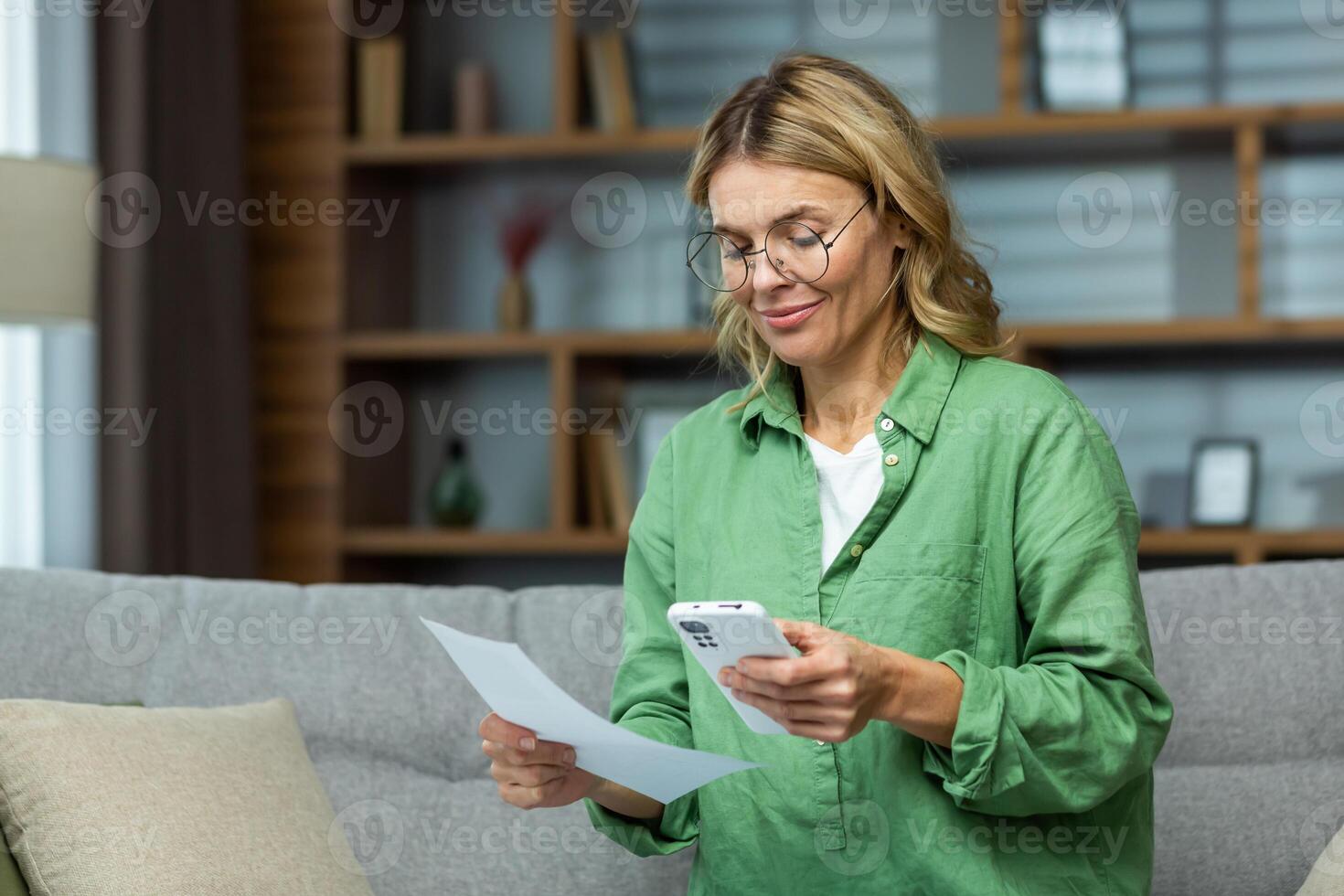 Mature experienced woman sitting on sofa at home, using app on smartphone to pay credit debts, smiling with satisfaction at ease and convenience of use. photo