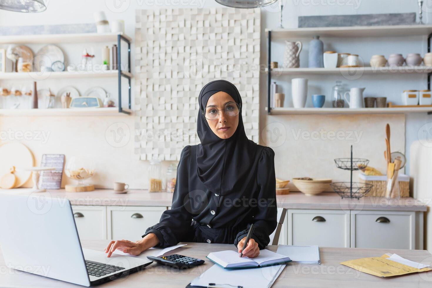 Successful and confident young woman in black hijab and burqa sitting at home at kitchen table and working remotely with documents and laptop. He looks seriously into the camera. photo
