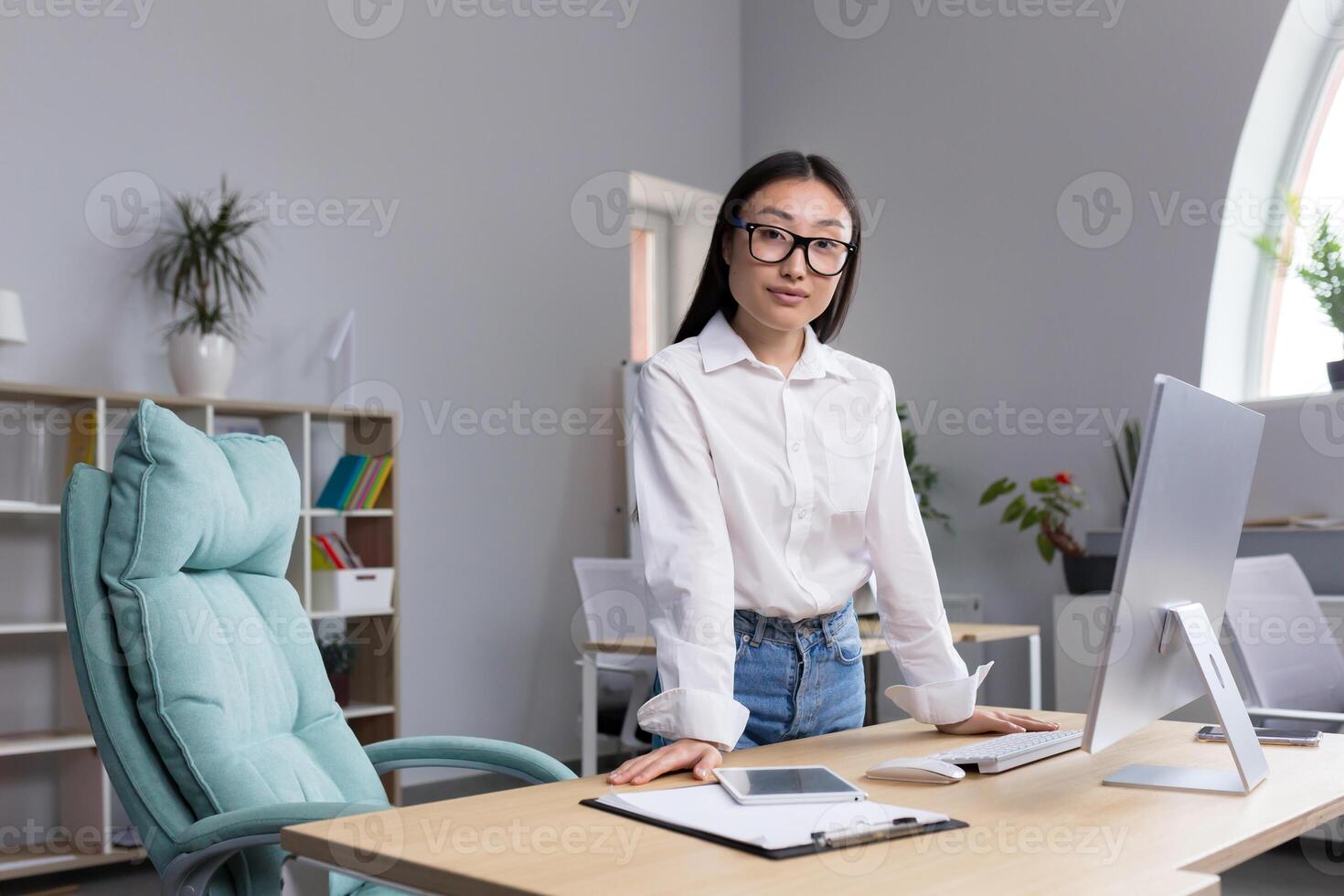 Portrait of a young beautiful Asian woman in glasses businesswoman, manager, director, boss standing in the office at the desk. She leans on the table with her hands, looks at the camera, smiles. photo