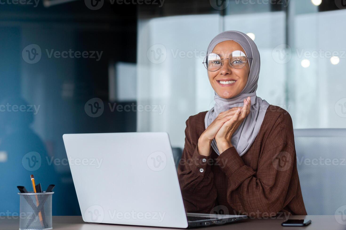Portrait of a young beautiful Arab female student in a hijab. Sitting at the table in the office with a laptop, looking at the camera, smiling, folded his hands together. photo