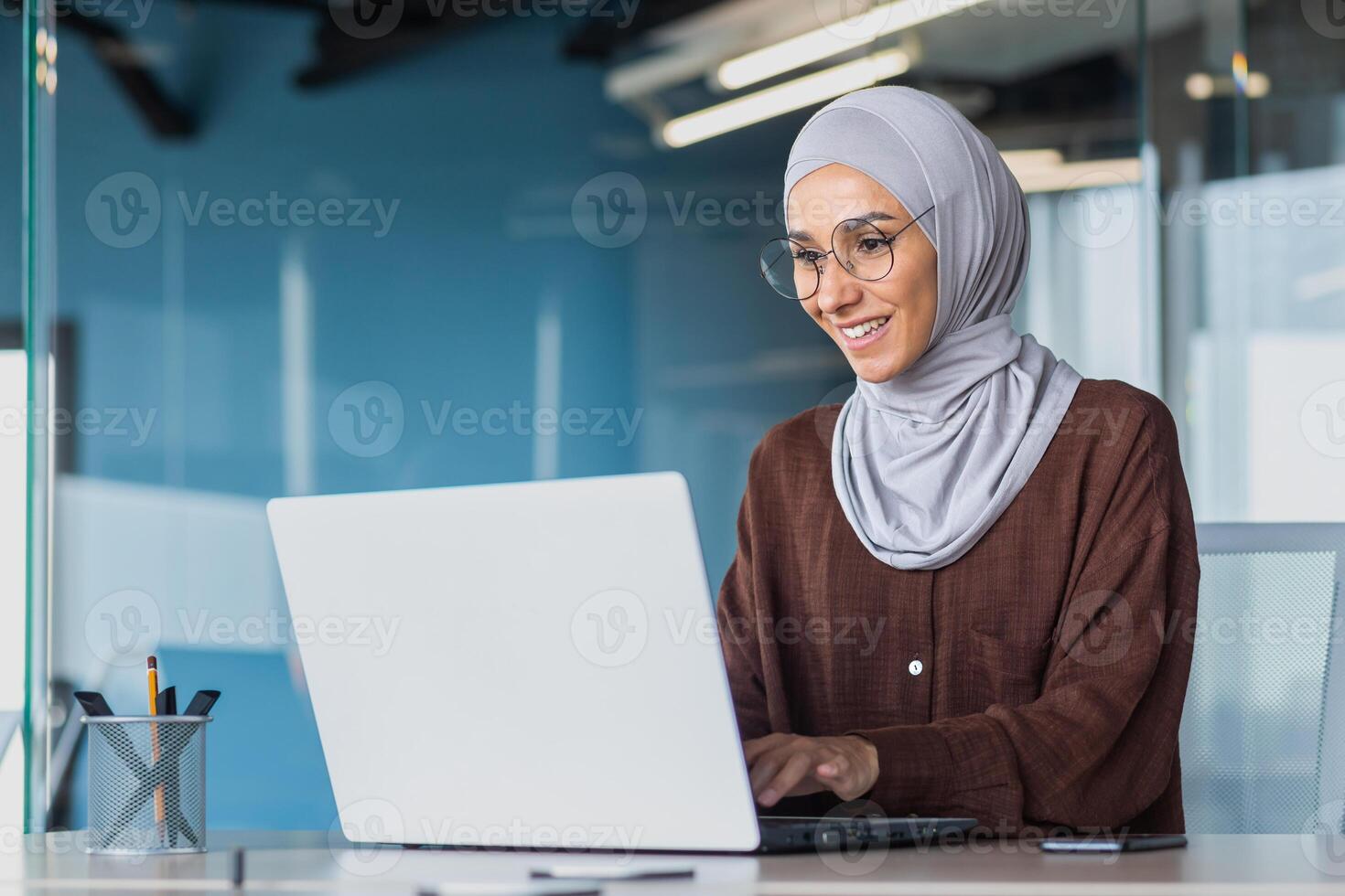 Happy and successful business woman in hijab working with laptop inside modern office building, muslim woman in glasses smiling and happy with work achievement. photo