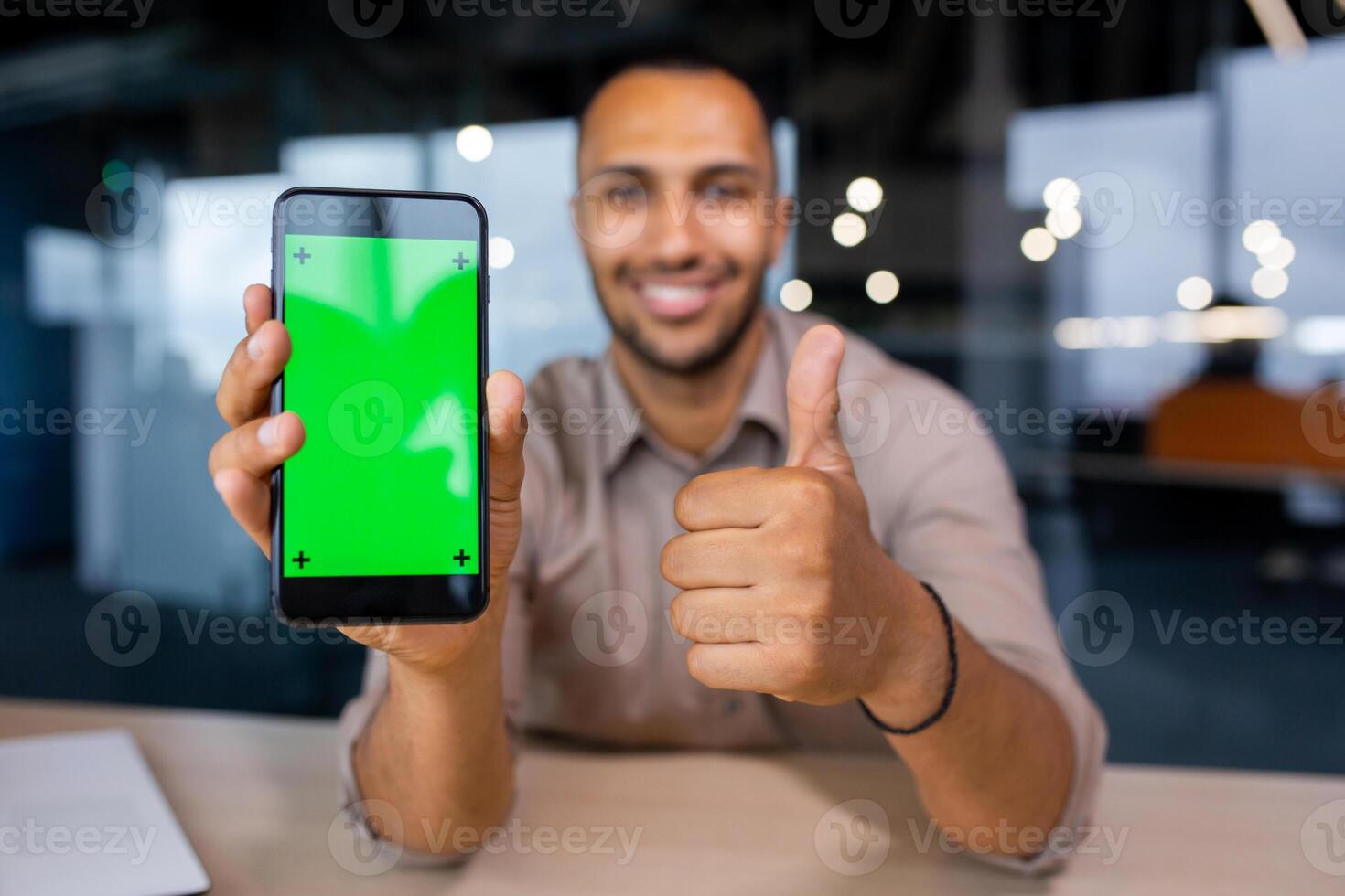 Happy man sitting in office holding phone with green screen and showing thumb up, phone in foreground. photo