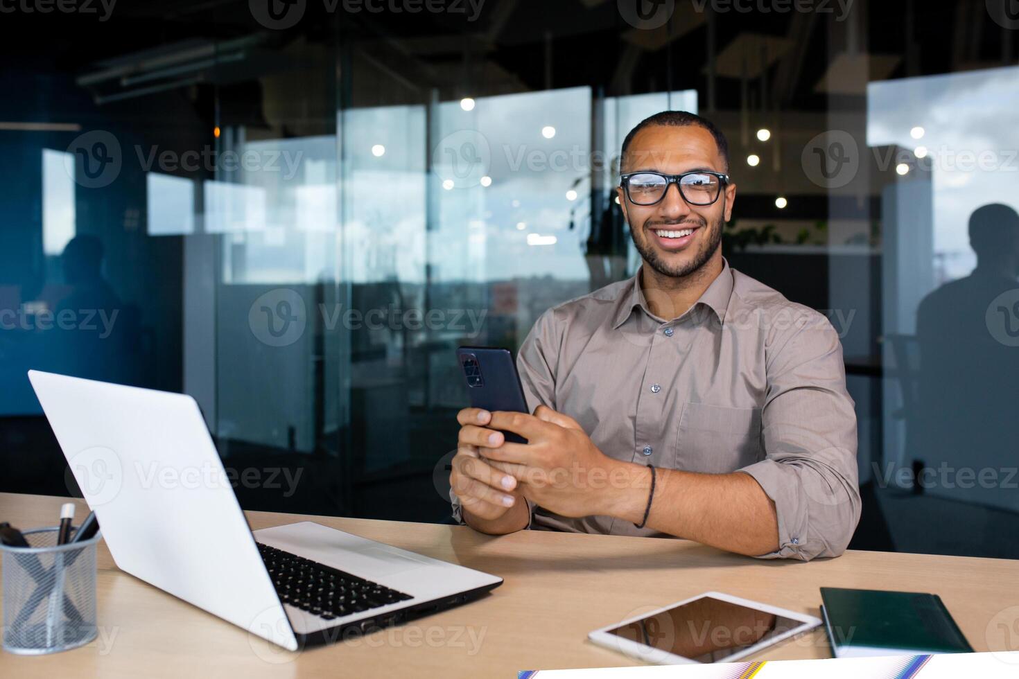 Portrait of a young successful businessman, the man is smiling and looking at the camera, holding a phone in his hands, using an application on a smartphone, working inside the office with a laptop. photo