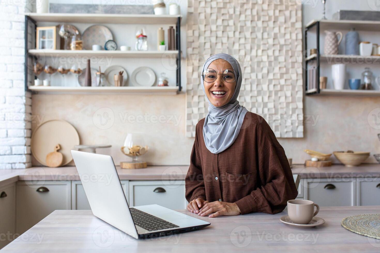 Portrait of young happy Muslim woman in hijab working and studying at home in kitchen using laptop. Smiling, she looks at the camera. photo