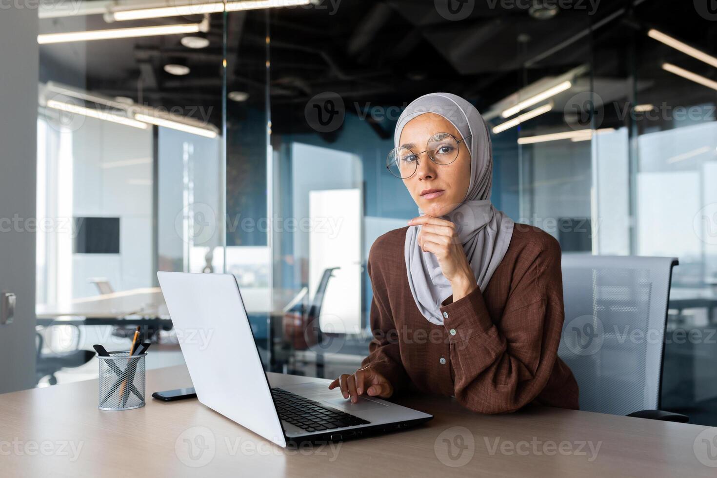 Portrait of a young woman Arab female student in hijab sitting in the office at the table and studying online at the laptop. He looks seriously at the camera. He holds his chin with his hand. photo
