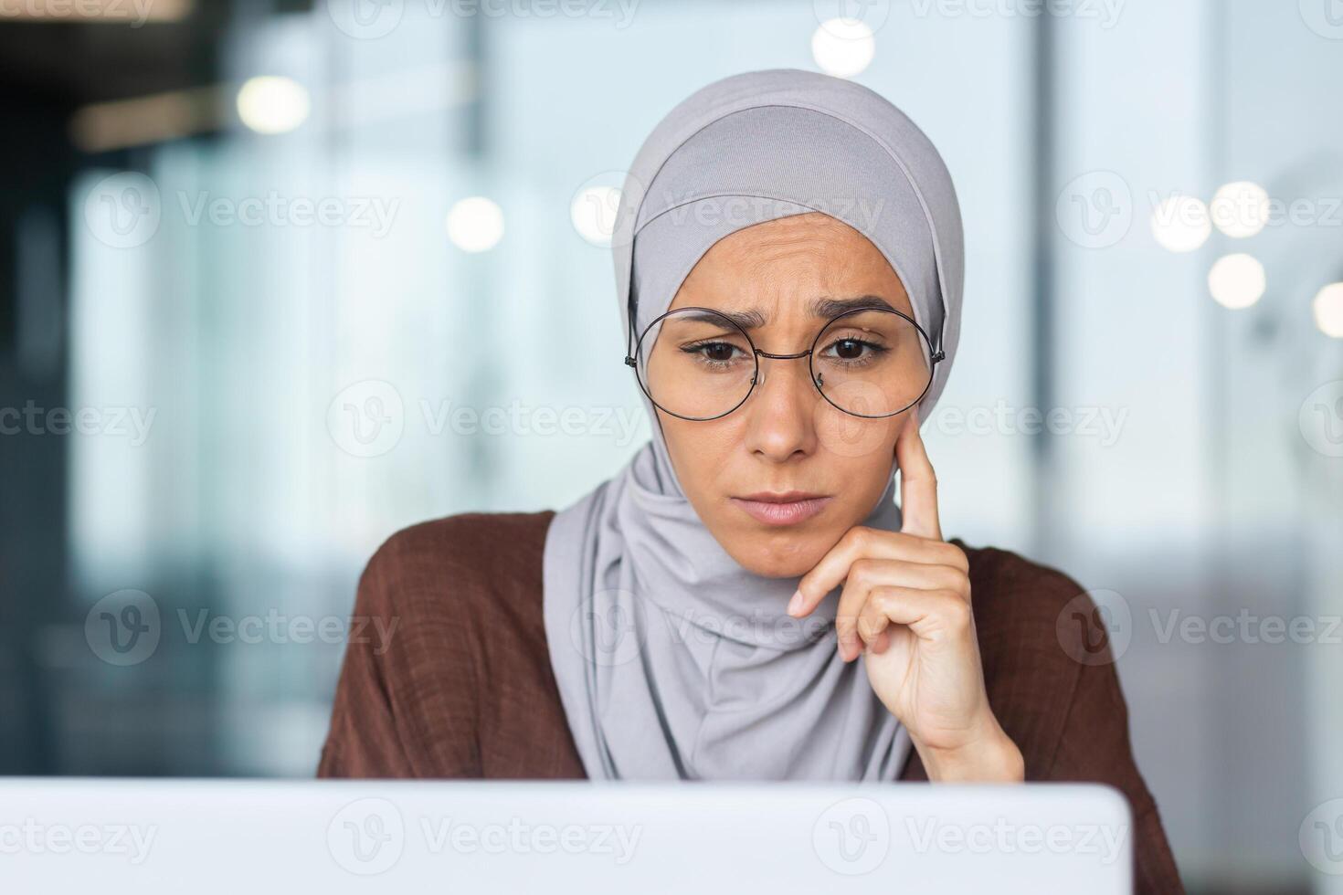 Close-up photo. Portrait of a young Arab woman in a hijab and glasses thoughtfully sitting in the office, working on a laptop. Leaning her head on her hand, she looks intently at the monitor. photo