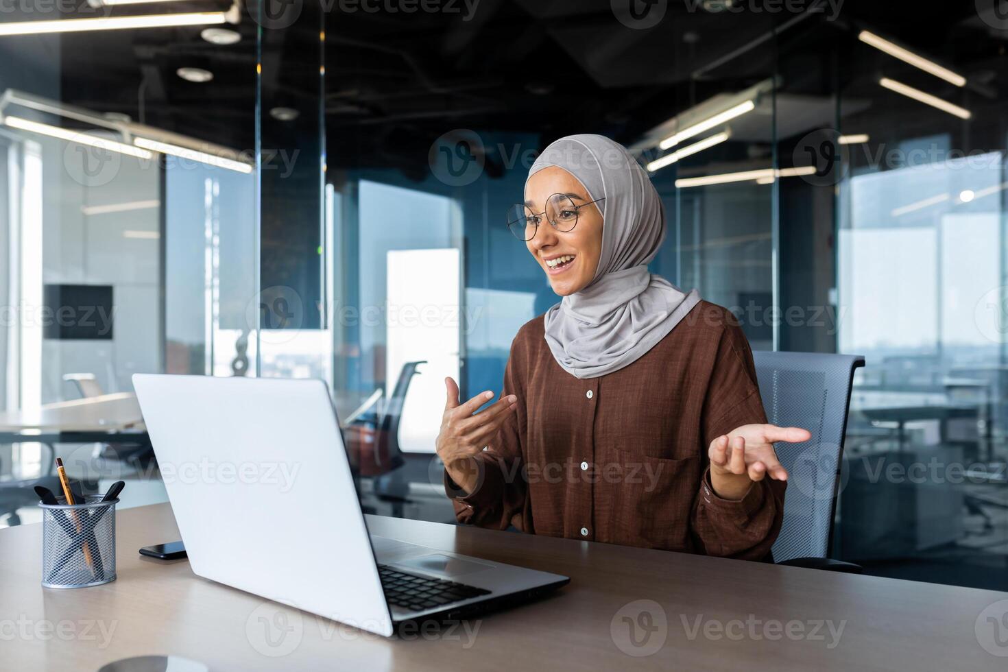 Successful and happy woman inside office at workplace, businesswoman boss in hijab talking on call using laptop sitting at table muslim woman joyfully gesturing to colleagues in online meeting photo
