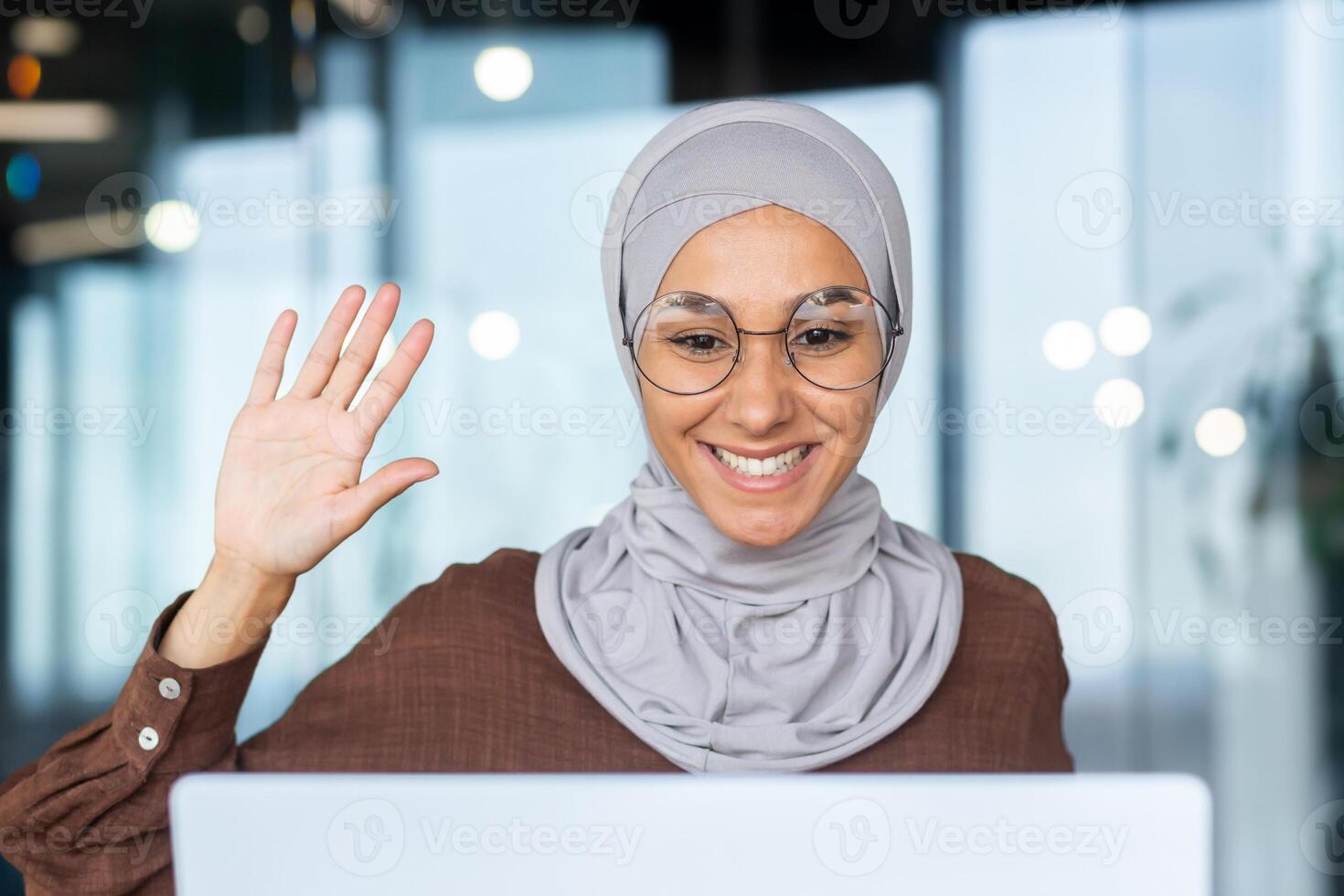 Young smiling muslim woman in hijab student talking on call via laptop. He waves his hand at the camera, says hello. photo