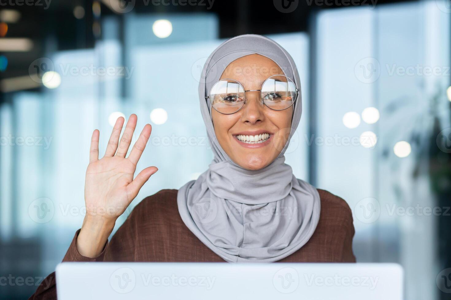 Close-up photo of a portrait of a young Muslim woman in a hijab working on a laptop and waving her hand to the camera with a smile.
