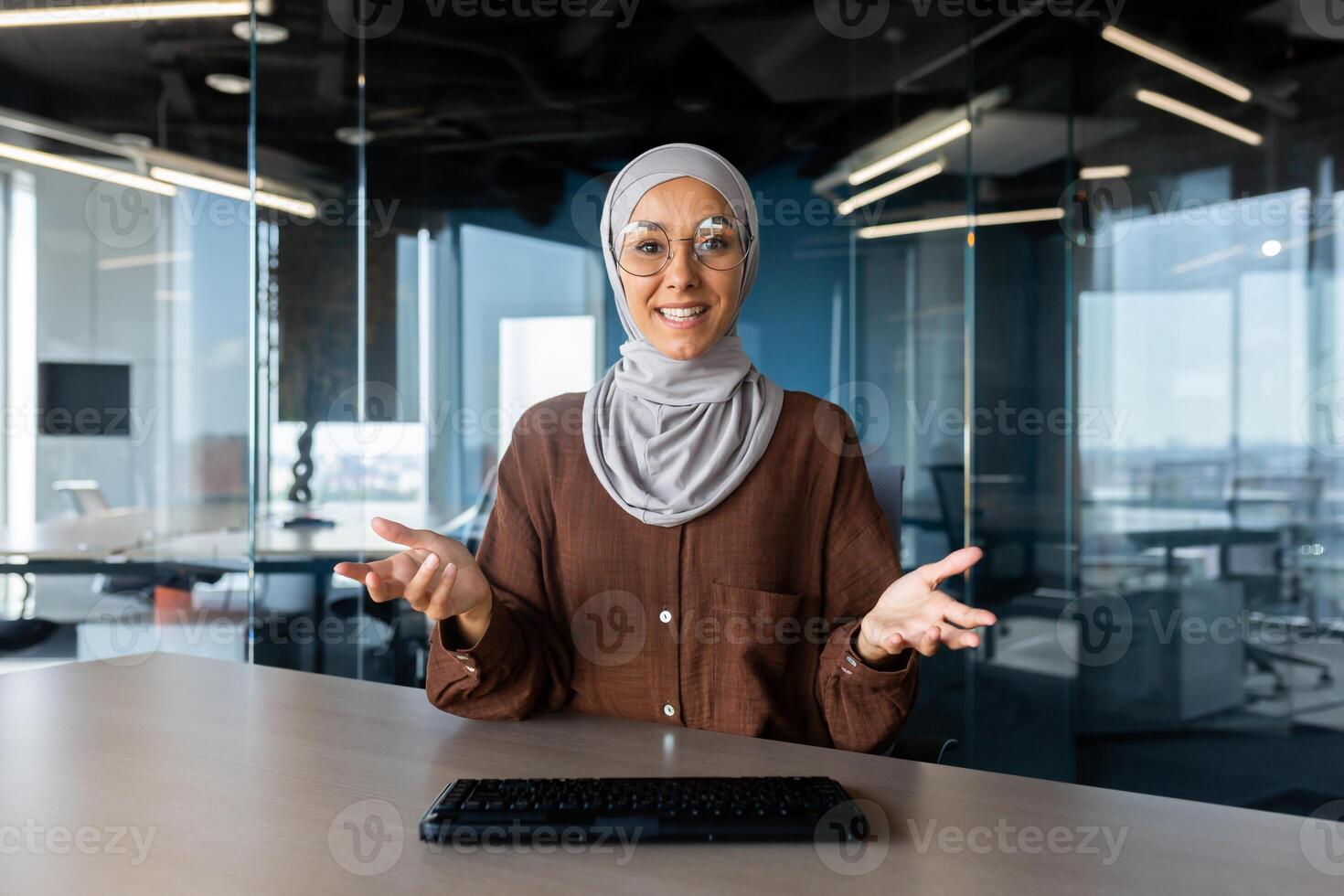 Close-up photo. Portrait of a smiling young Muslim business woman wearing a hijab and glasses sitting at the desk in the office and talking to the camera. photo