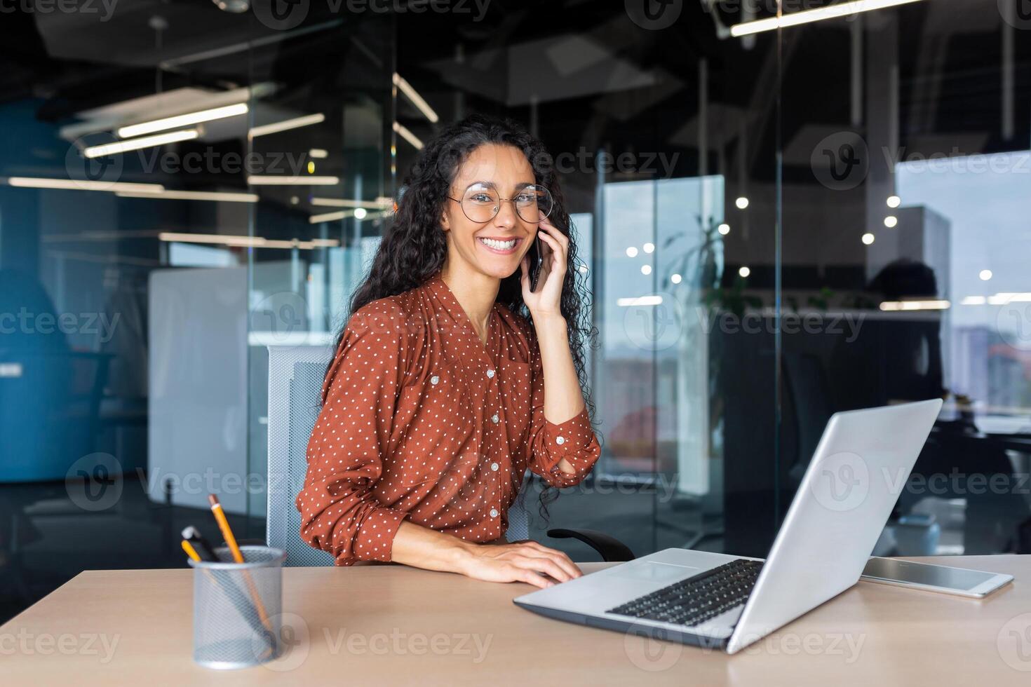 Portrait of successful latin american business woman, office worker smiling and talking on phone looking at camera inside office, accountant financier using laptop at work. photo