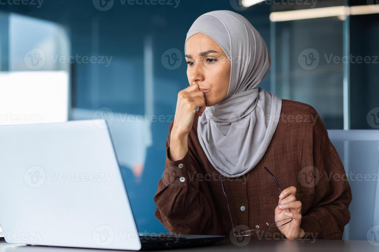 Portrait of a serious young woman in a hijab sitting in front of a laptop, holding glasses, thoughtfully and intently looking at the monitor. photo