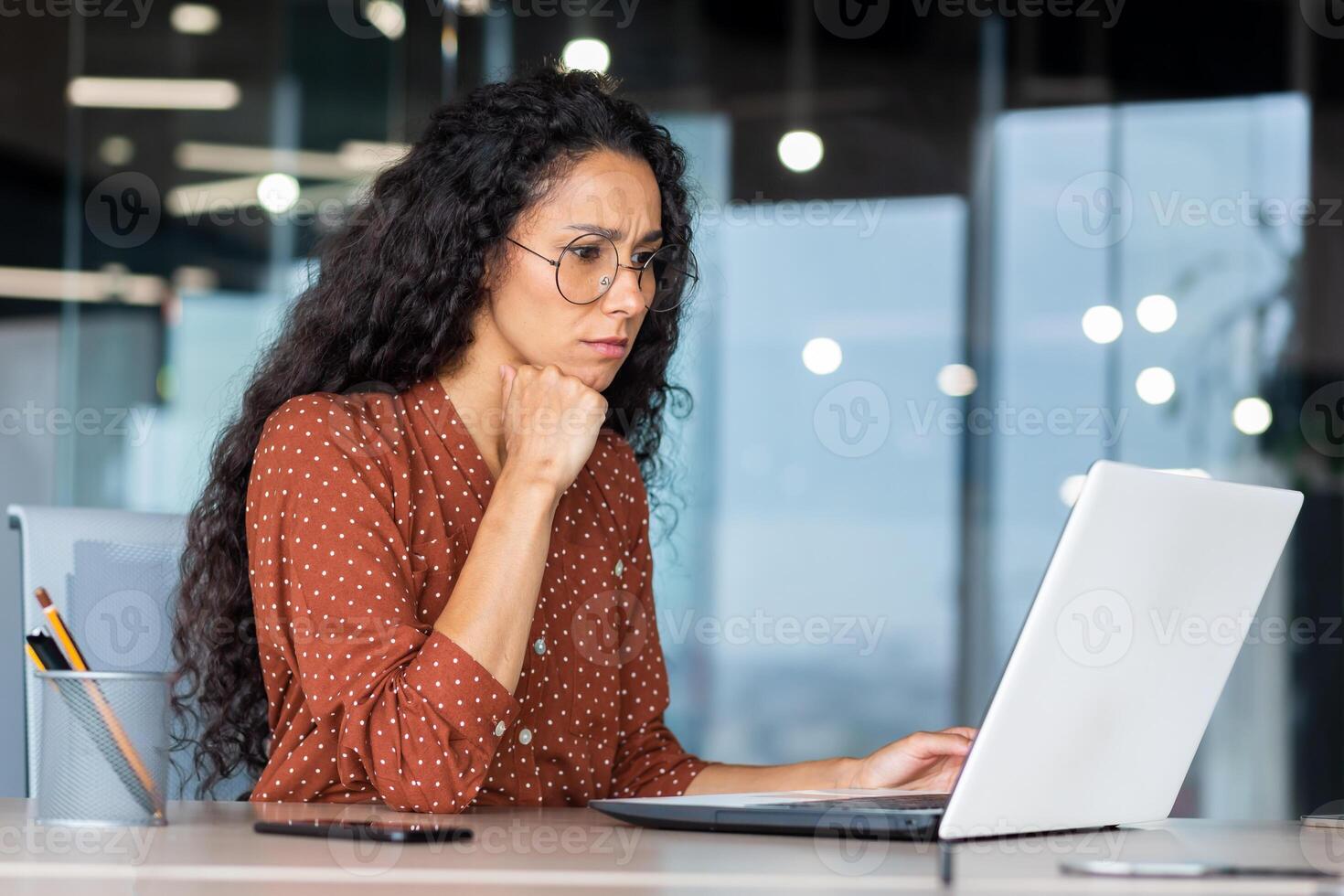 Serious thinking Hispanic businesswoman working inside modern office, female programmer looking at laptop testing new software. photo