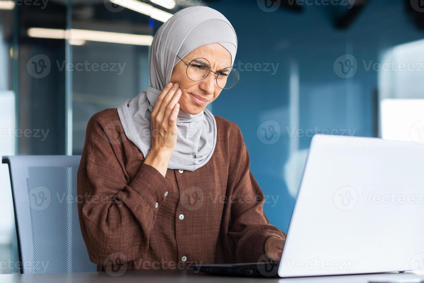 Close-up photo. Toothache at the workplace. Portrait of a young woman who works in the office at a table with a laptop, holding her cheek, doing a massage, feeling a strong toothache. photo