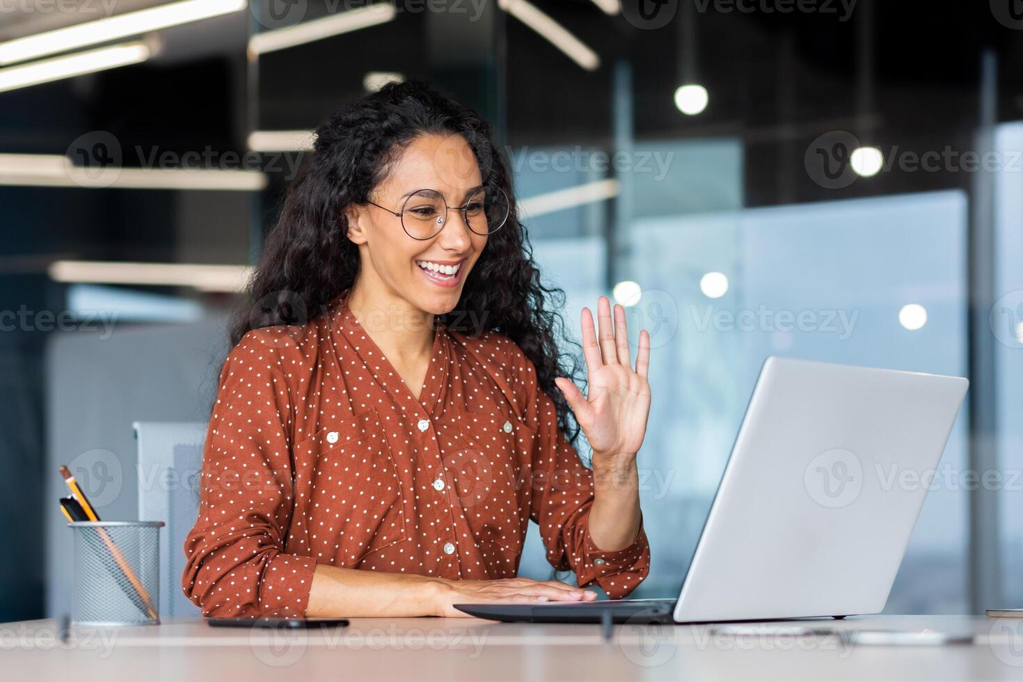 Successful and beautiful hispanic woman working inside modern office building, businesswoman using laptop for call smiling and waving, greeting gesture, online conference with colleagues. photo