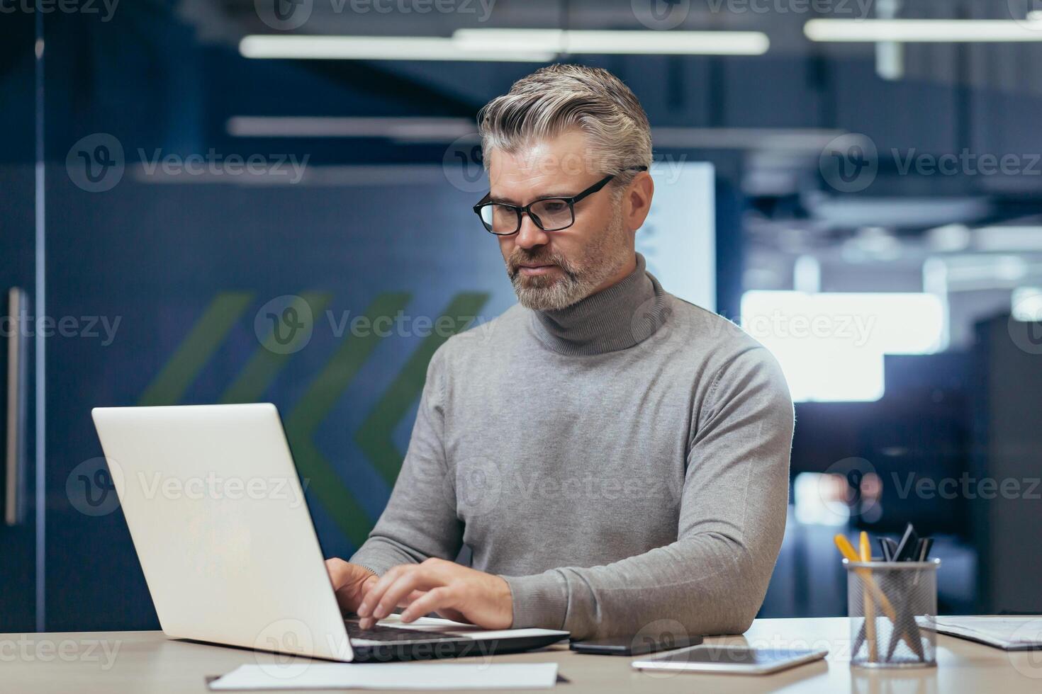 Portrait of a senior gray-haired man, a businessman, director, founder, who is concentrating on working in the office at a desk with a laptop. photo