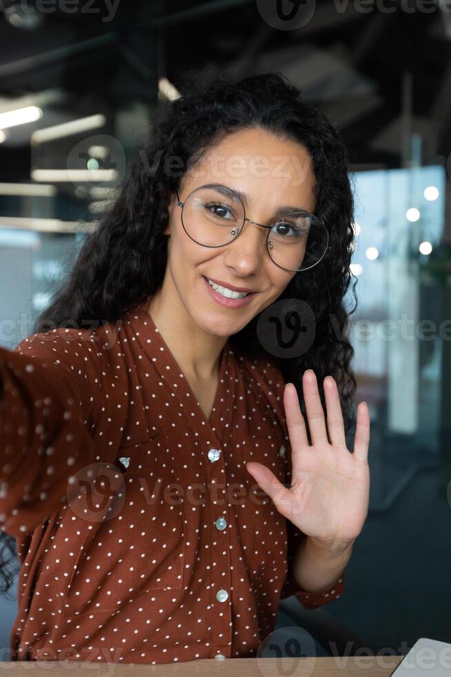 Vertical shot, young beautiful Hispanic business woman with curly hair talking on call, waving at smartphone camera, using remote communication app photo