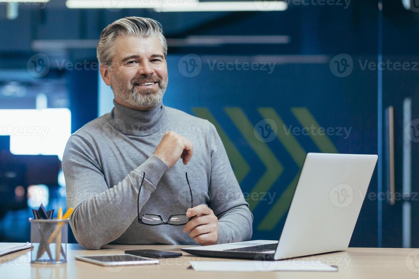 Portrait of senior gray-haired businessman, experienced mature man working with laptop inside office building, investor smiling and looking at camera photo