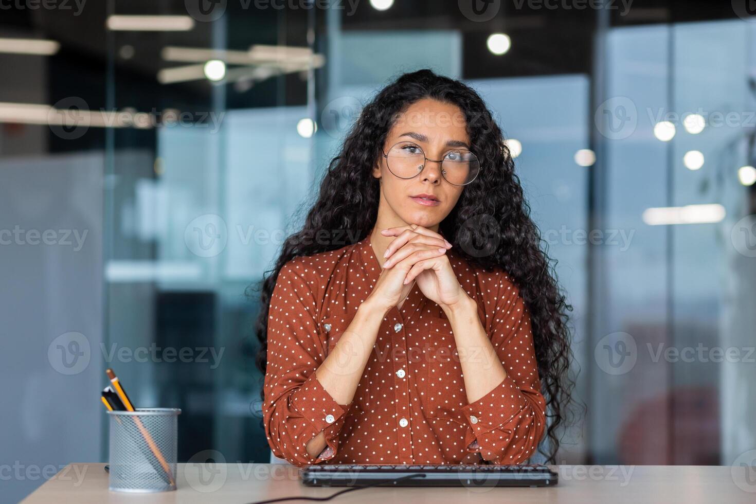 Serious Latin American business woman listening to a call report, thoughtfully looking at the web camera, female employee working inside the office of an online customer service support tech. photo