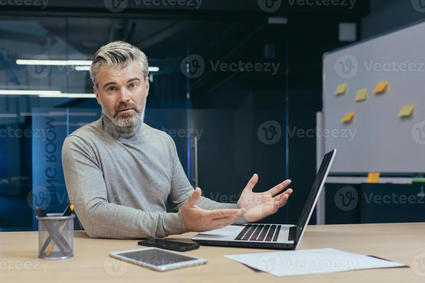 Portrait of upset gray-haired mature businessman, senior man looking at camera upset, businessman working in middle of office building using laptop photo