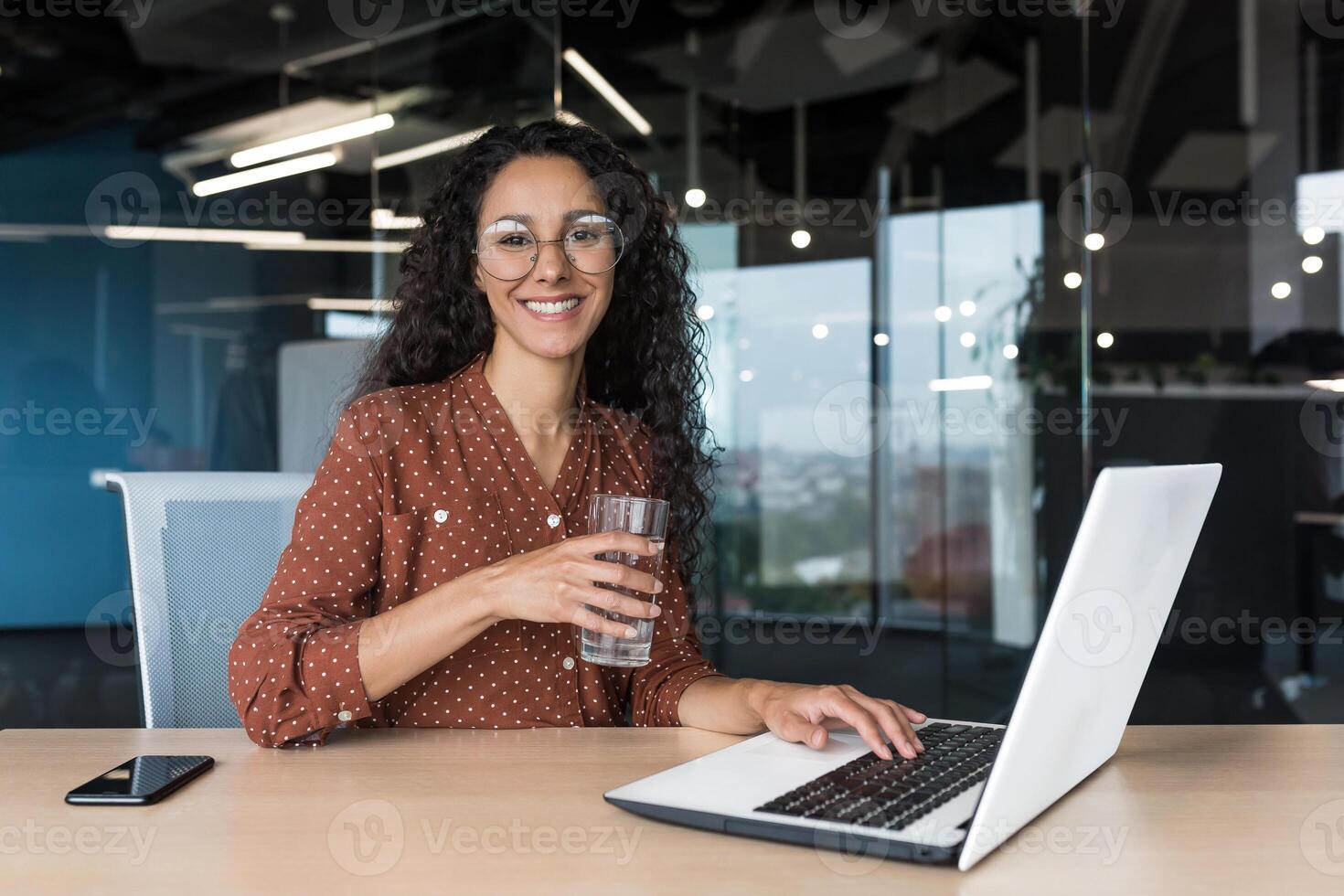 Portrait of happy and successful business woman inside modern office building, female worker smiling and looking at camera, holding glass of clean water, working on laptop at desk photo