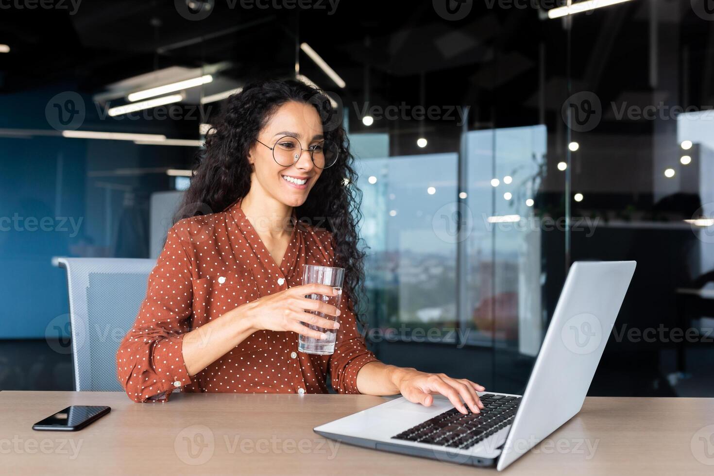 Successful and happy Latin American business woman at work inside the office, female worker holds a glass of clean water in her hands and drinks, businesswoman is satisfied achievement, uses a laptop. photo