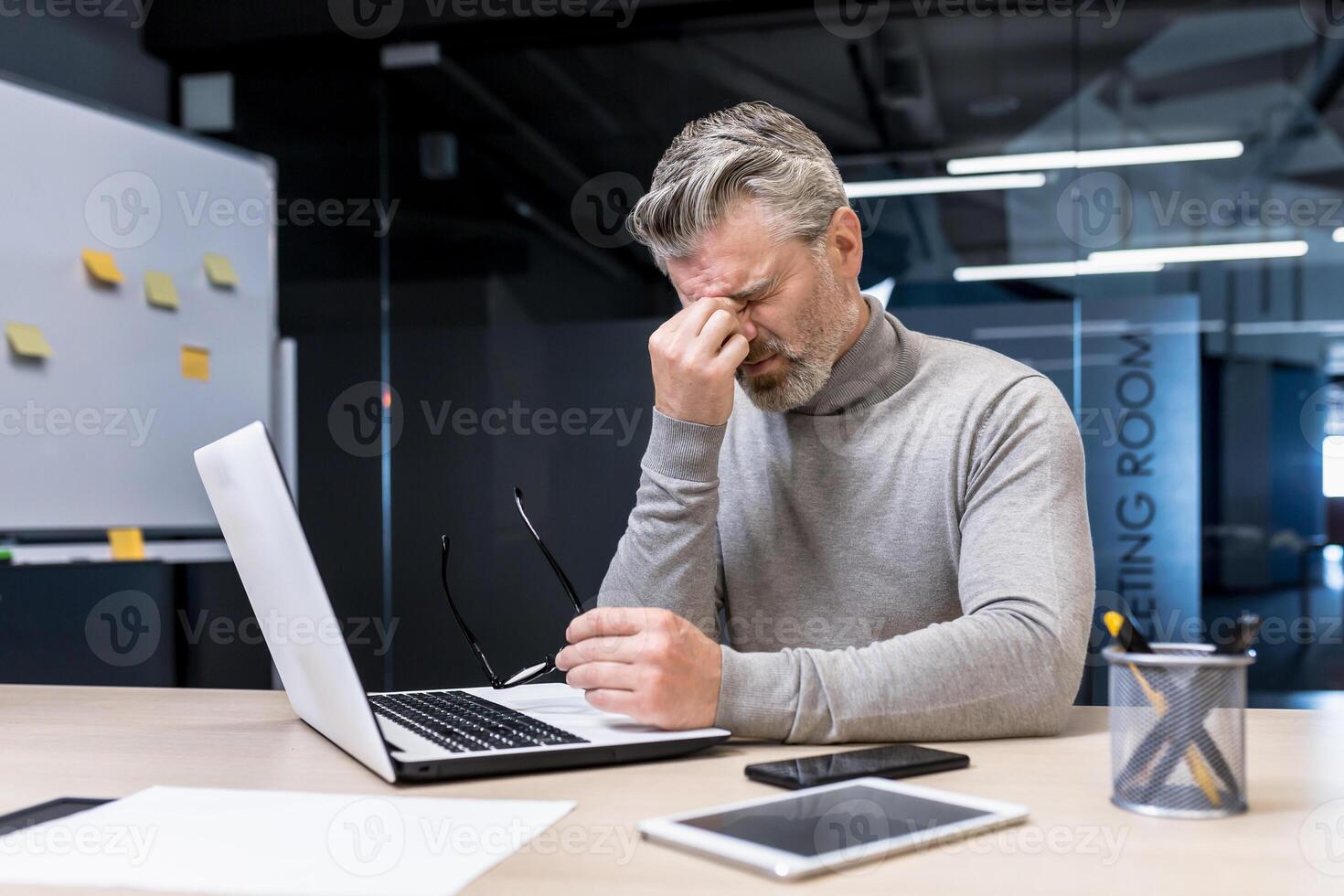 Stressful, tense, tired handsome male businessman sitting in office at desk with laptop and phone. Takes a break, took off his glasses, covered his eyes with his hand. photo