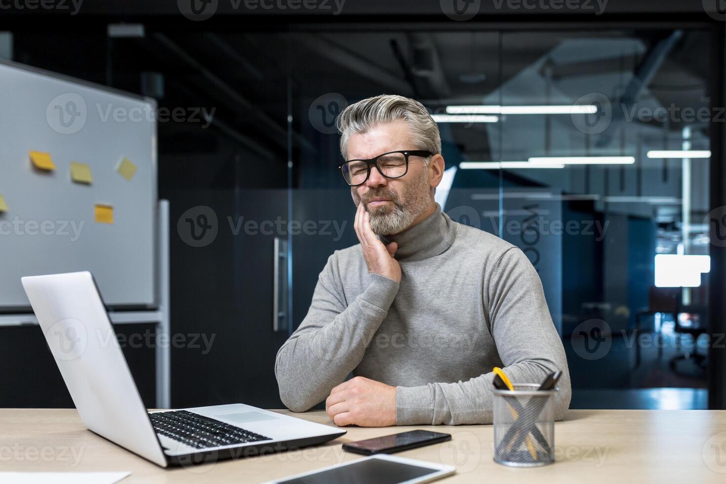 Serious toothache in mature gray-haired office worker, senior experienced businessman working inside office building using laptop at desk, man holding toothache. photo