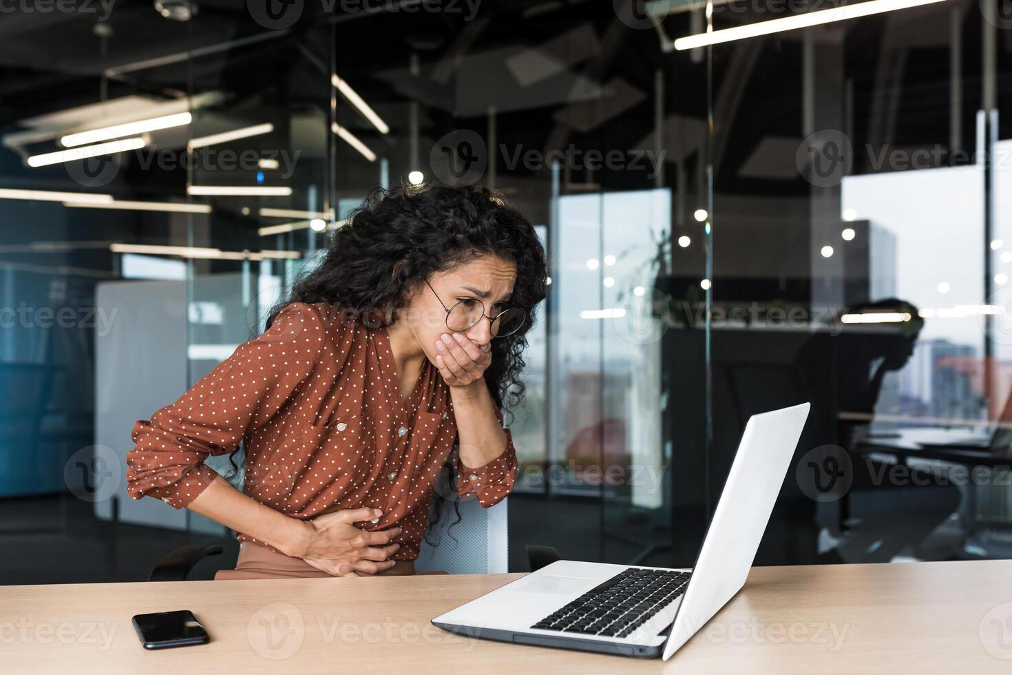 Sick business woman working at work, having severe nausea and abdominal pain, latin american working inside office building using laptop. photo